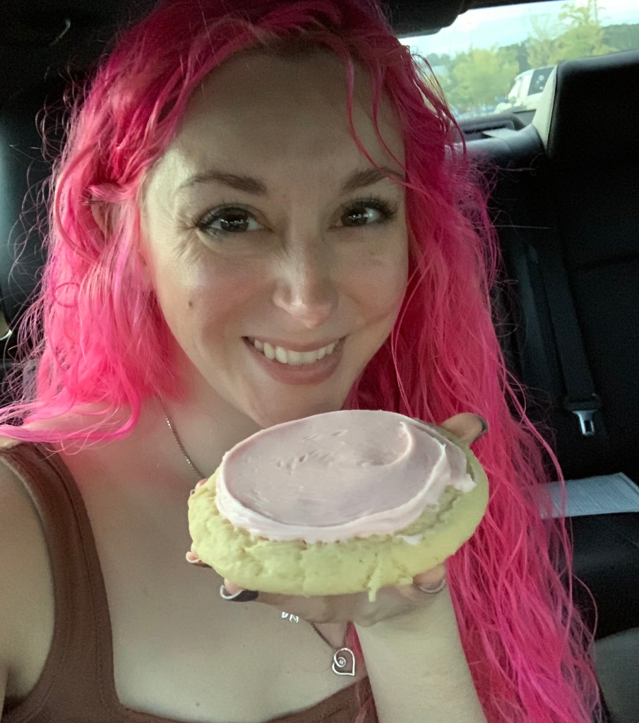 Amanda Nichols in a car, smiling with bright pink hair, holding a large pink-frosted cookie.
