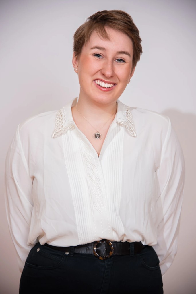 Delaney Gilmour, studio portrait with short brown hair, white lace-collar blouse, smiling against a light grey background.